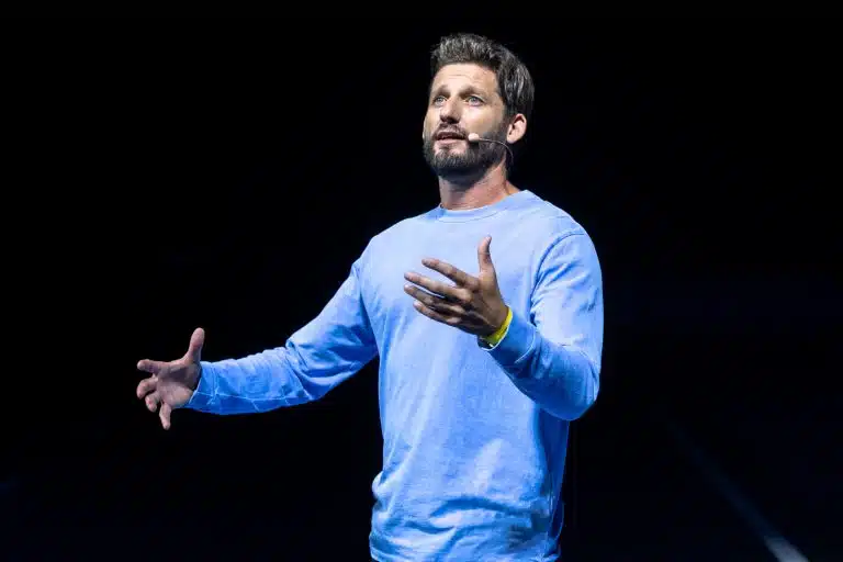 Man wearing a blue shirt speaking passionately on stage with a headset microphone, lit dramatically against a dark background during a live presentation or keynote