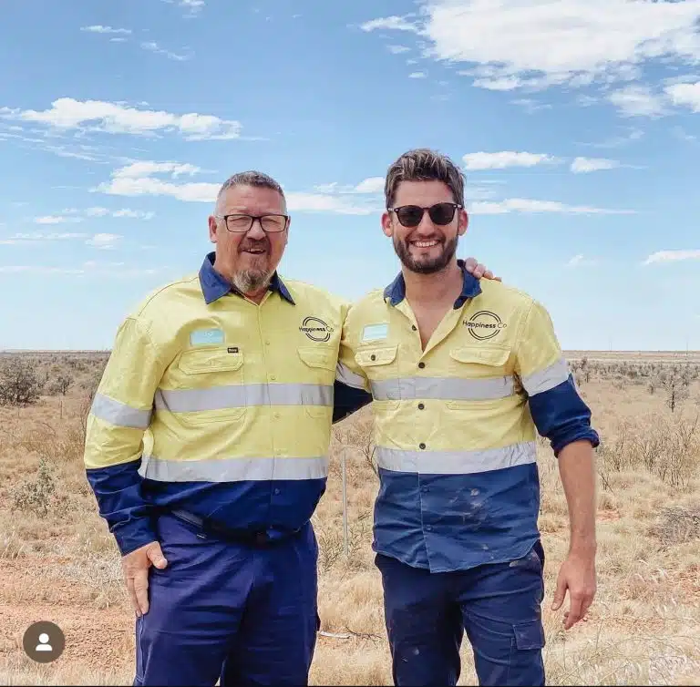 Two men wearing high-visibility Happiness Co uniforms standing side by side and smiling in an outback landscape under a bright blue sky with scattered clouds
