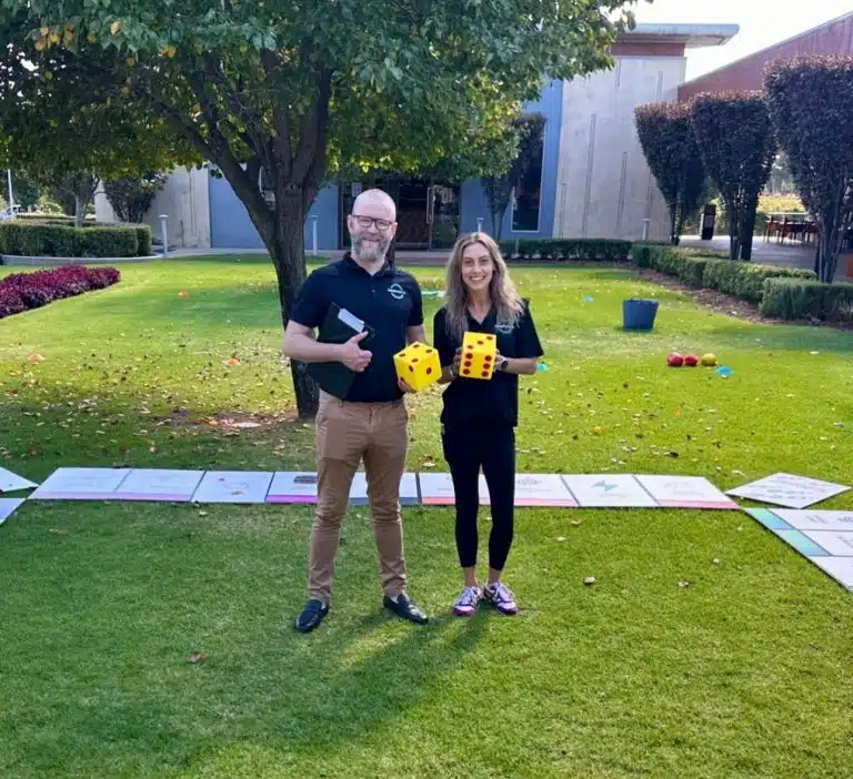 Two facilitators from Happiness Co standing on a grassy lawn during an outdoor wellbeing activity, holding large foam dice with a life-sized board game spread across the grass.