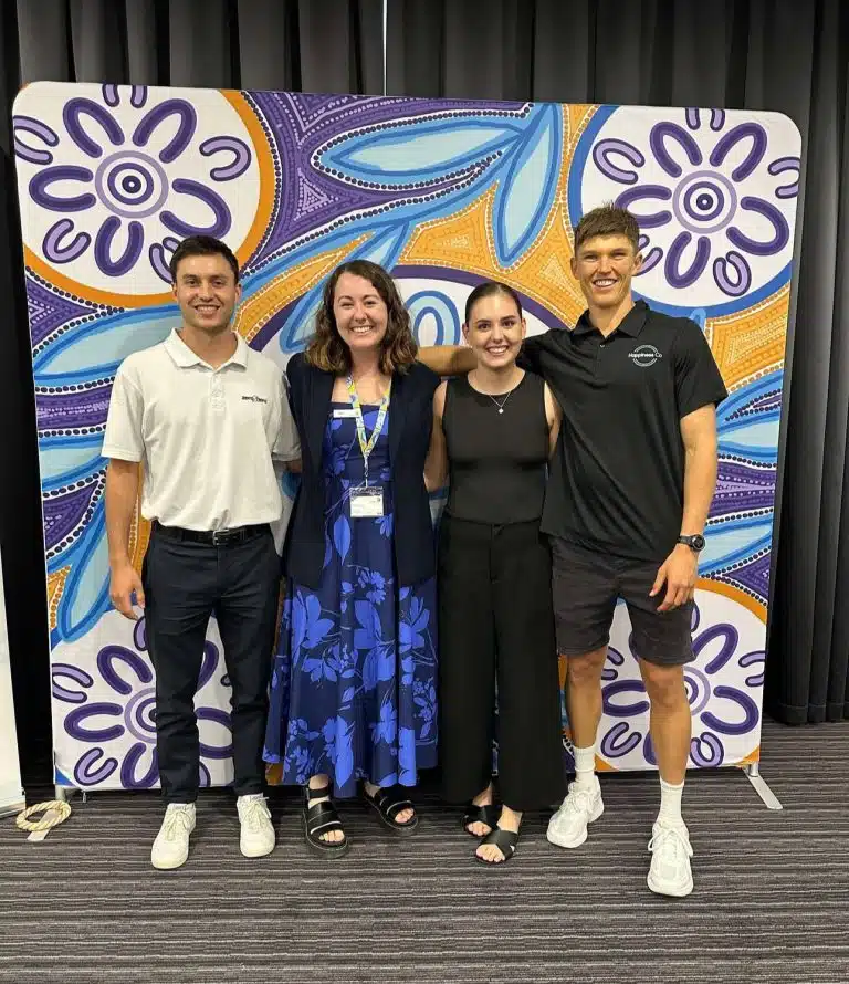 Four young professionals smiling and standing in front of a vibrant Aboriginal artwork backdrop at an indoor community or education event.