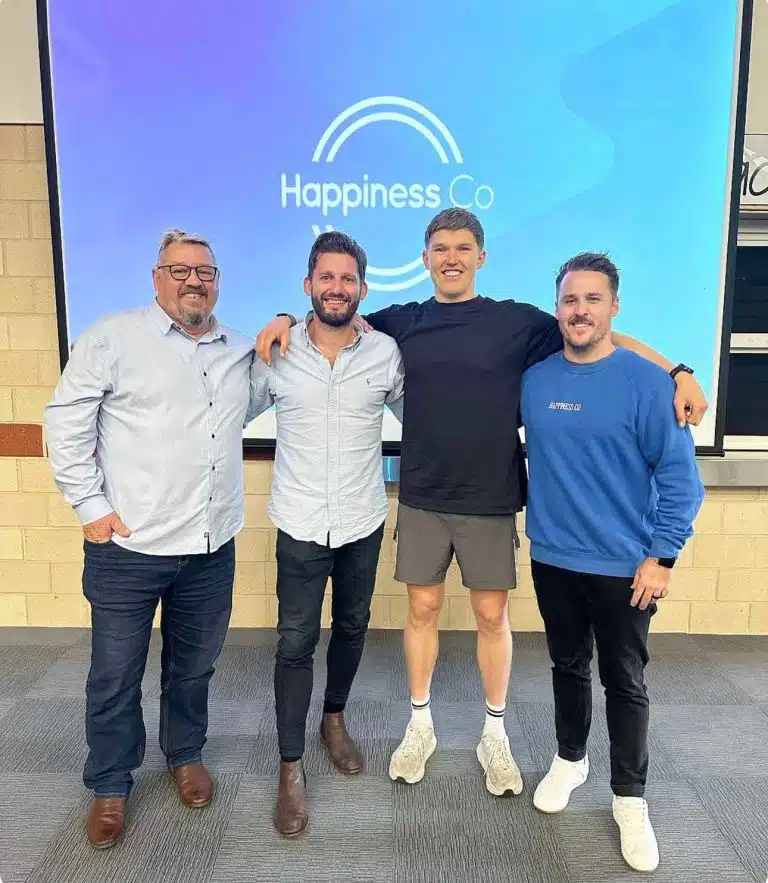 Four men standing together and smiling in front of a screen displaying the Happiness Co logo at an indoor event or presentation.
