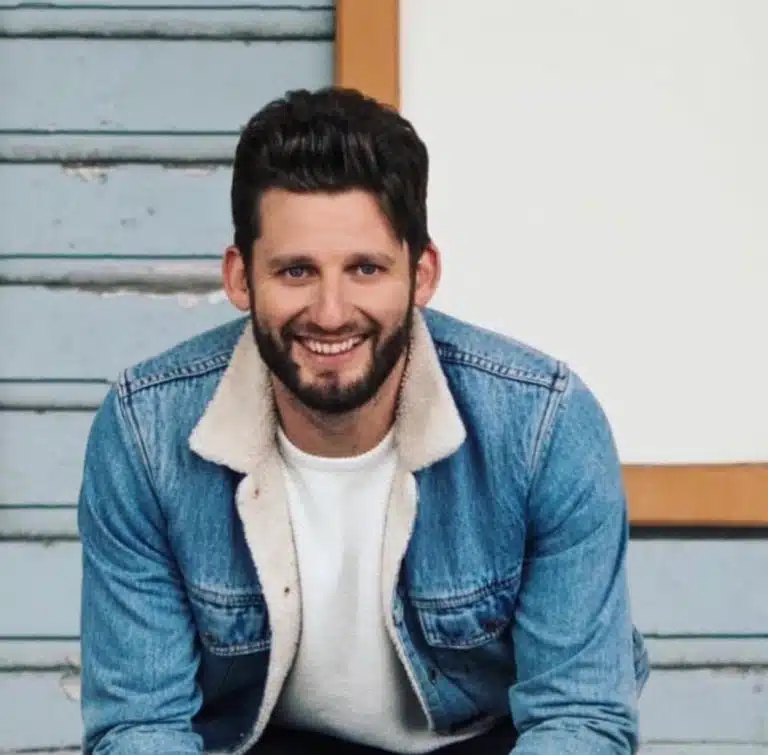 Smiling man in a denim jacket with a shearling collar sitting outdoors on wooden steps in a relaxed pose